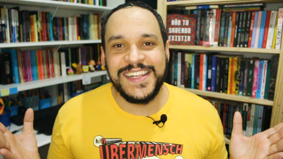 Ao fundo, prateleiras com livros. À frente, o Bruno sorrindo, de barba curta, camisa amalera com a palavra Ubermensch escrita em vermelho.